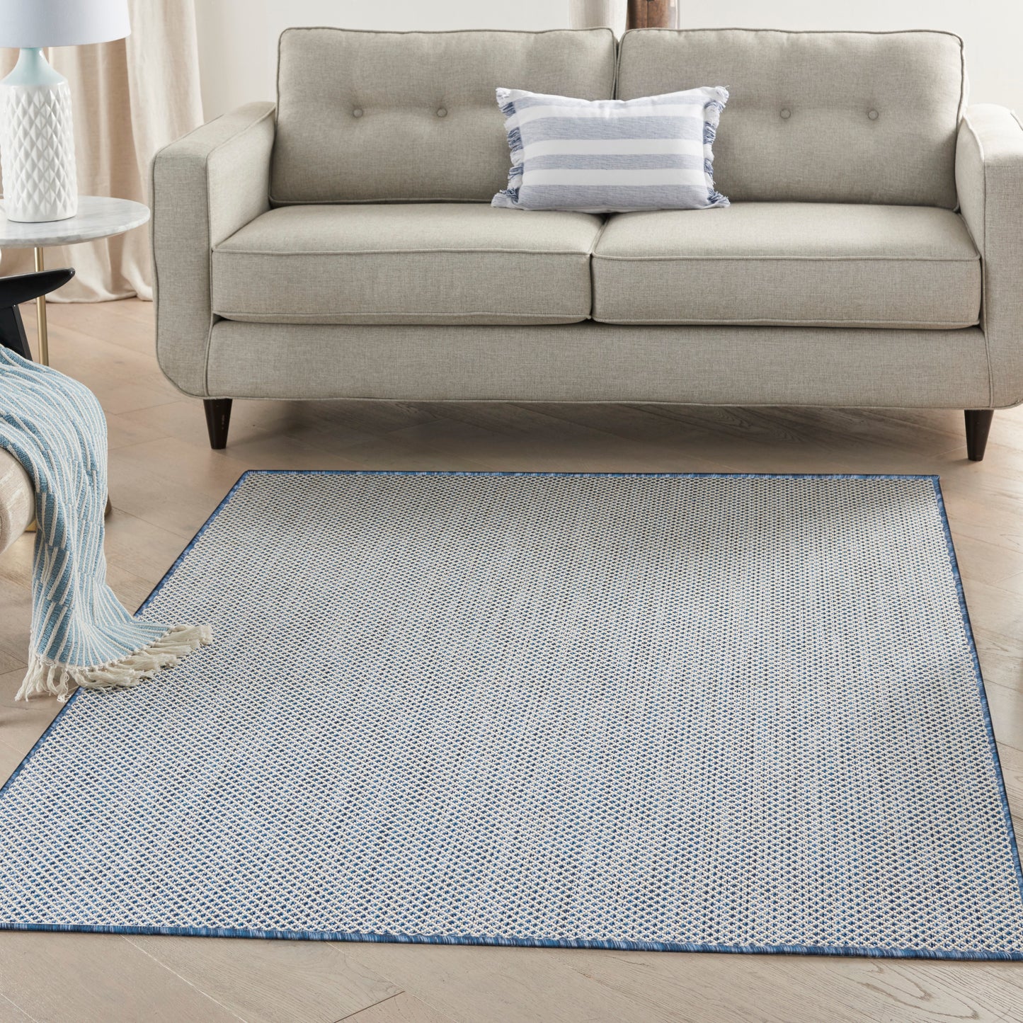 Beige fabric mid-century sofa with tufted back, blue striped pillow, and a light blue woven area rug with darker blue border.