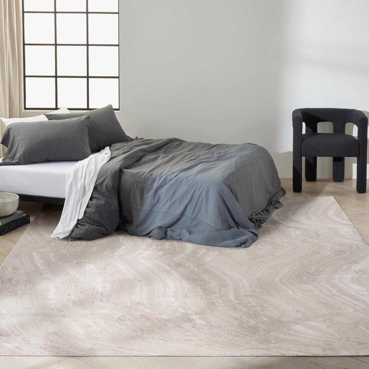 Modern bedroom with a low bed in gray bedding, white sheet, and a large pale gray rug; black accent chair by a grid-window.