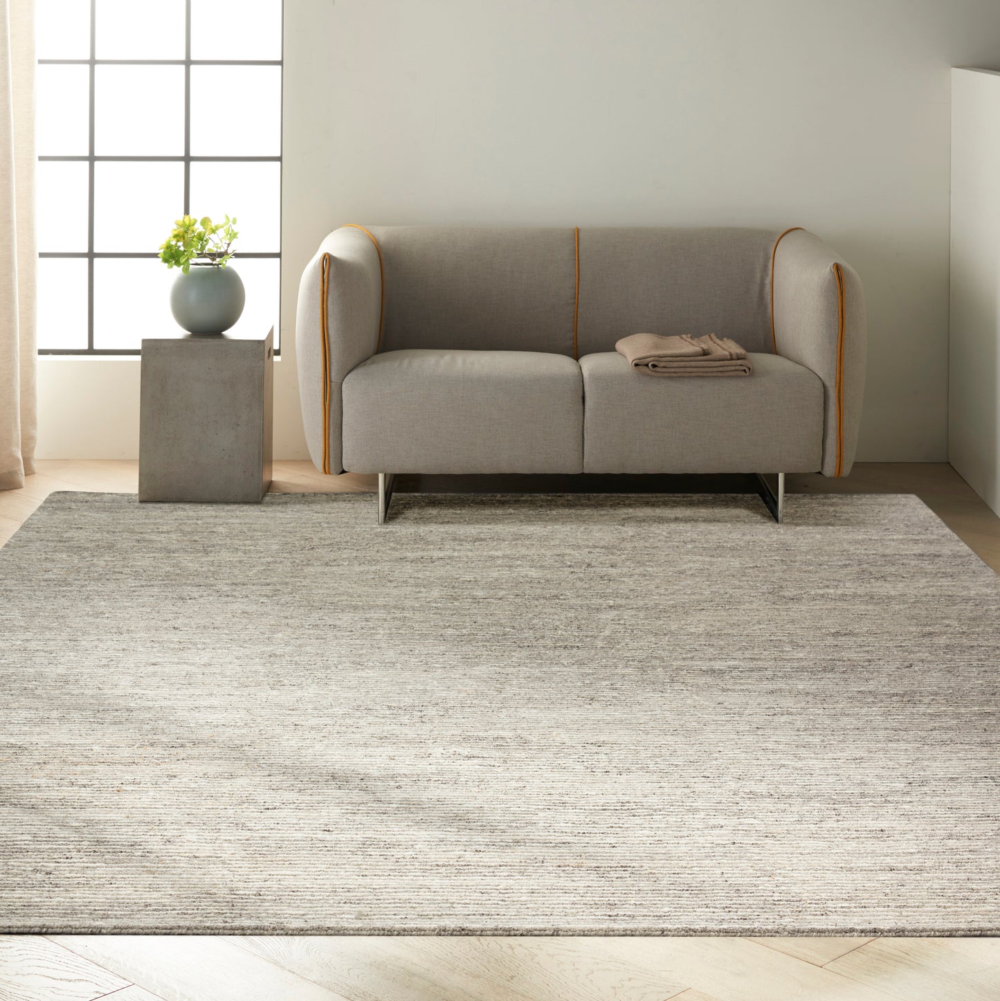 Light gray large area rug with subtle texture; a light gray two-seater sofa with orange piping on rounded arms and a beige throw sits on the rug.