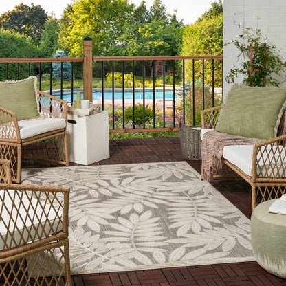 Leaf-pattern outdoor rug on a wooden deck, with outdoor wicker seating featuring beige cushions & green pillows; pool & greenery beyond.