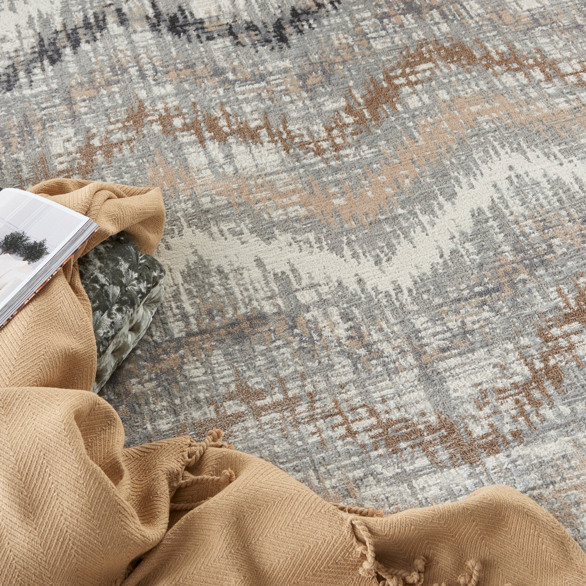 Rectangular area rug, 5' x 7', in gray, beige & brown with an abstract pattern, paired with a tan throw blanket & an open magazine.