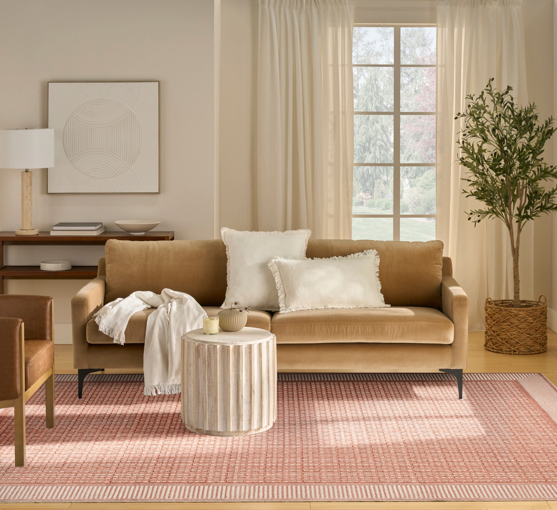 Tan fabric sofa with white pillows and beige throw, round wood coffee table, pink-patterned rug, potted tree by window.