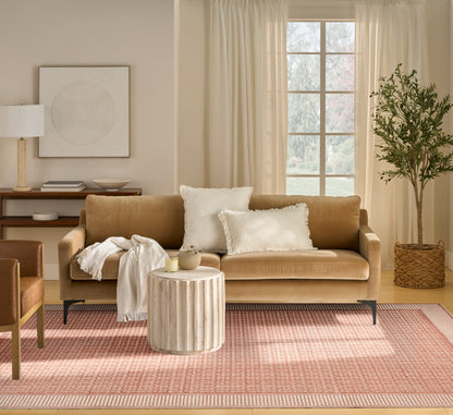 Tan fabric sofa with white pillows and beige throw, round wood coffee table, pink-patterned rug, potted tree by window.