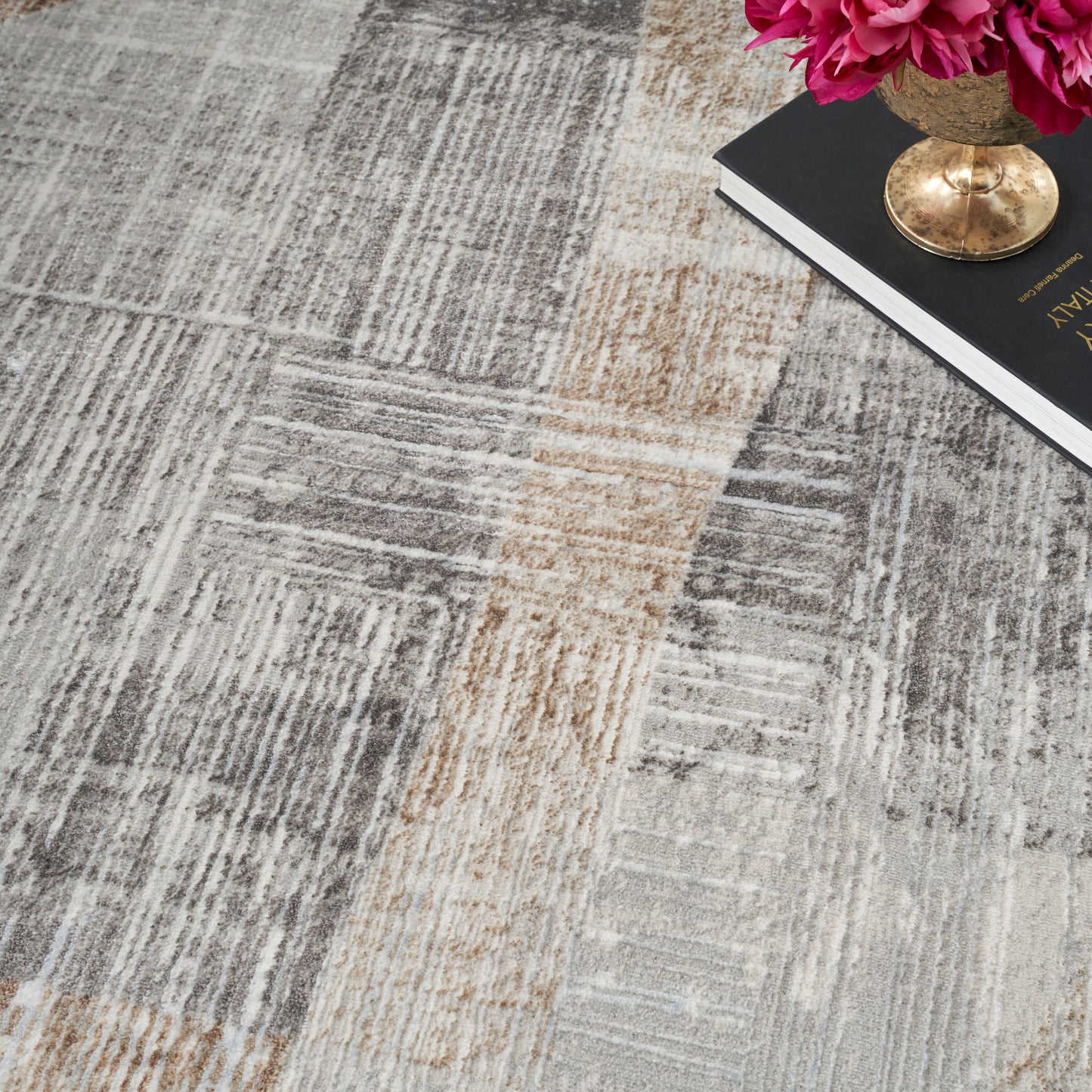 Modern patchwork area rug in gray and beige tones; low-pile woven texture, shown with pink flowers and a gold vase.