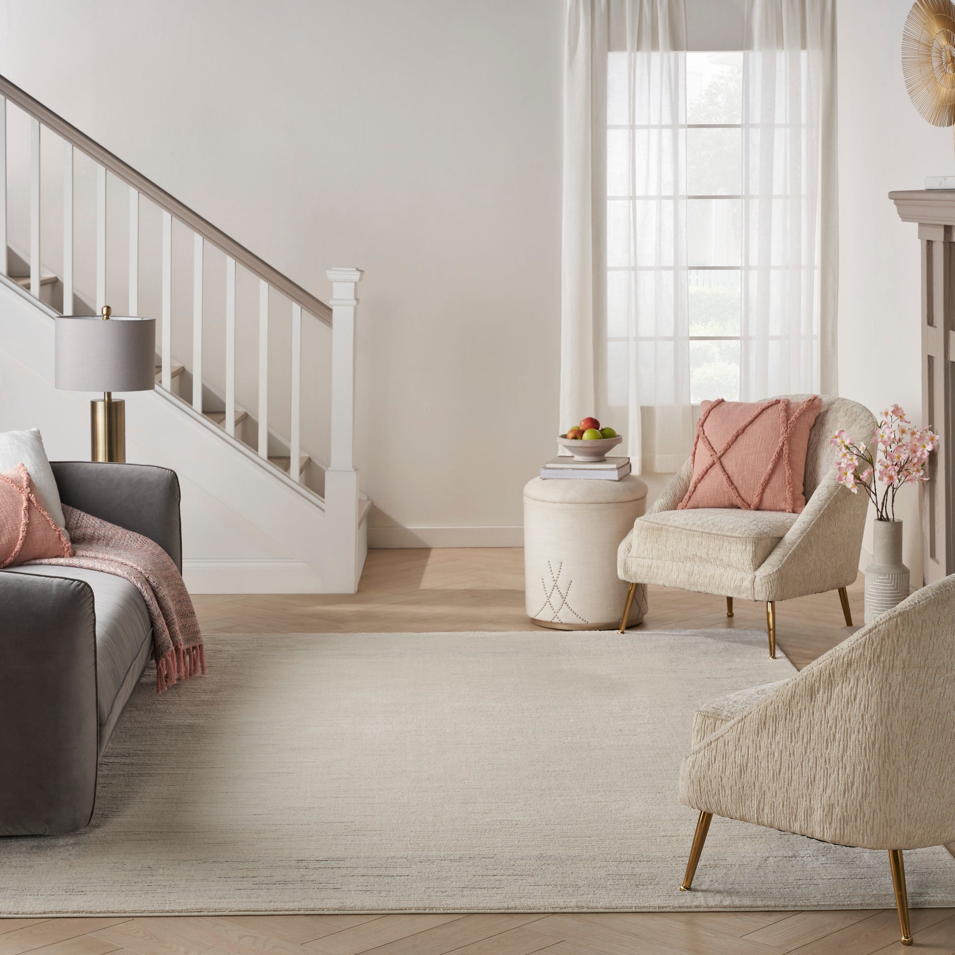 Bright neutral living room with gray sofa, pink throw pillows, beige accent chair, light rug, staircase, sheer curtains.