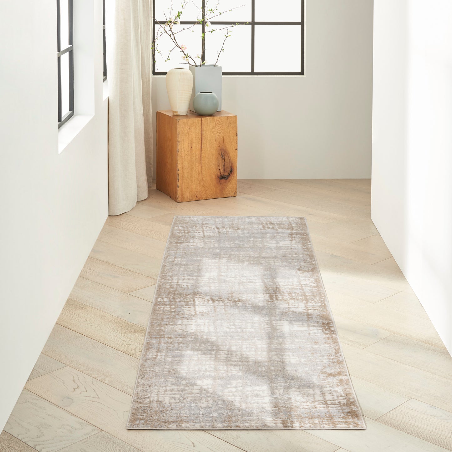 Bright hallway with light wood floor, beige-gray patterned runner rug, and ceramic vases on a wooden pedestal by a window.