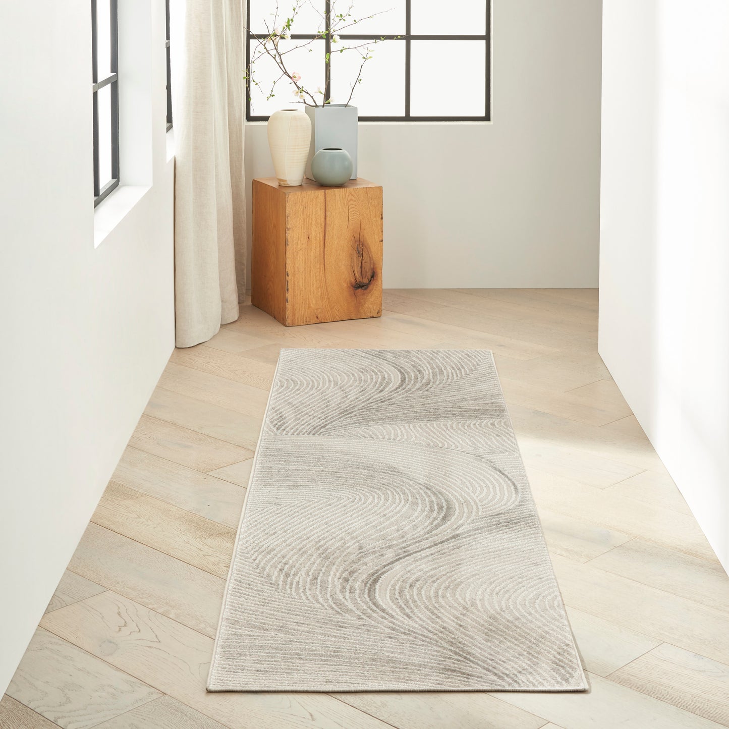 Long light gray rug runner with subtle circular wave pattern on pale wood floor in bright hallway by a window.