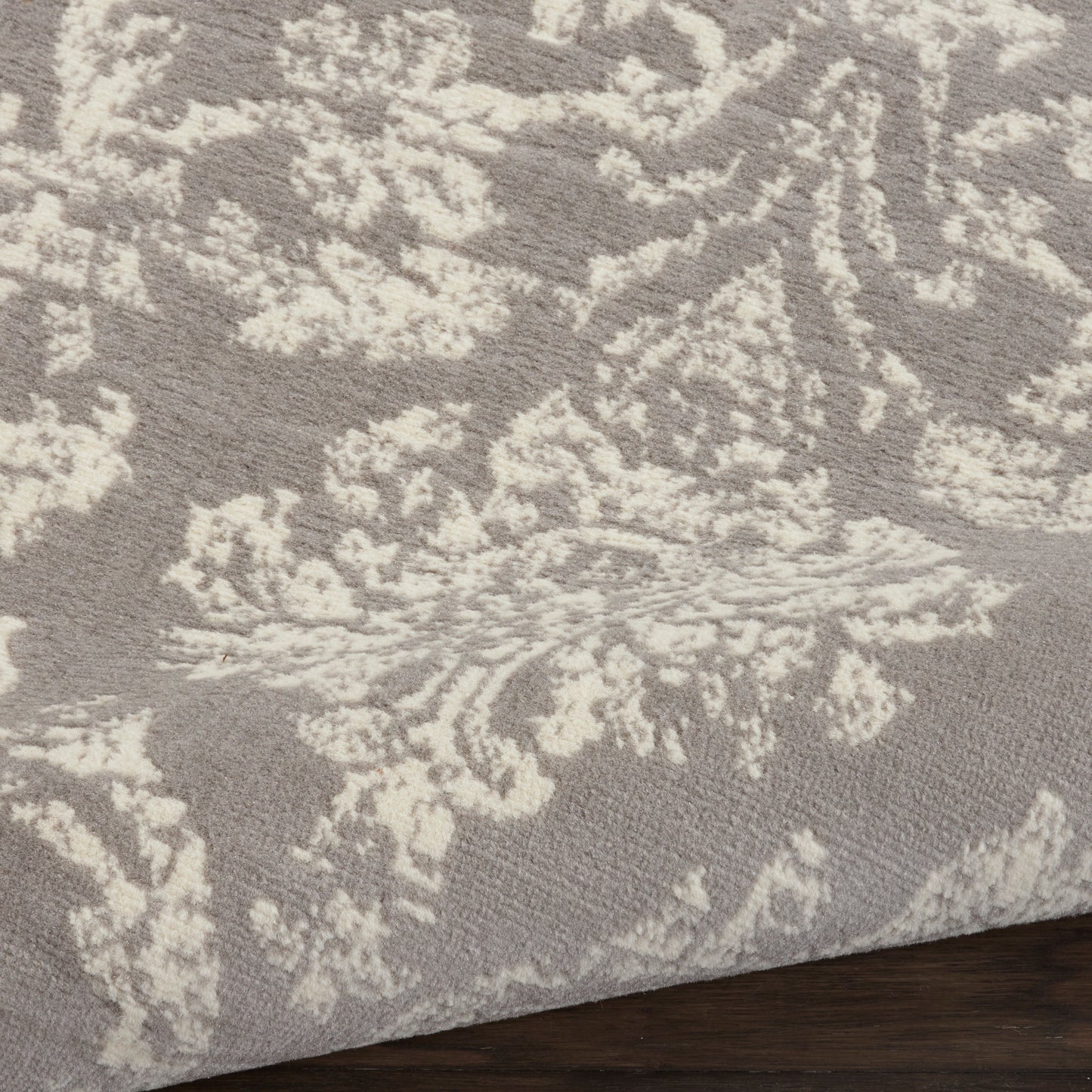 Taupe and cream damask patterned area rug with soft pile, edge folded on dark wood floor.