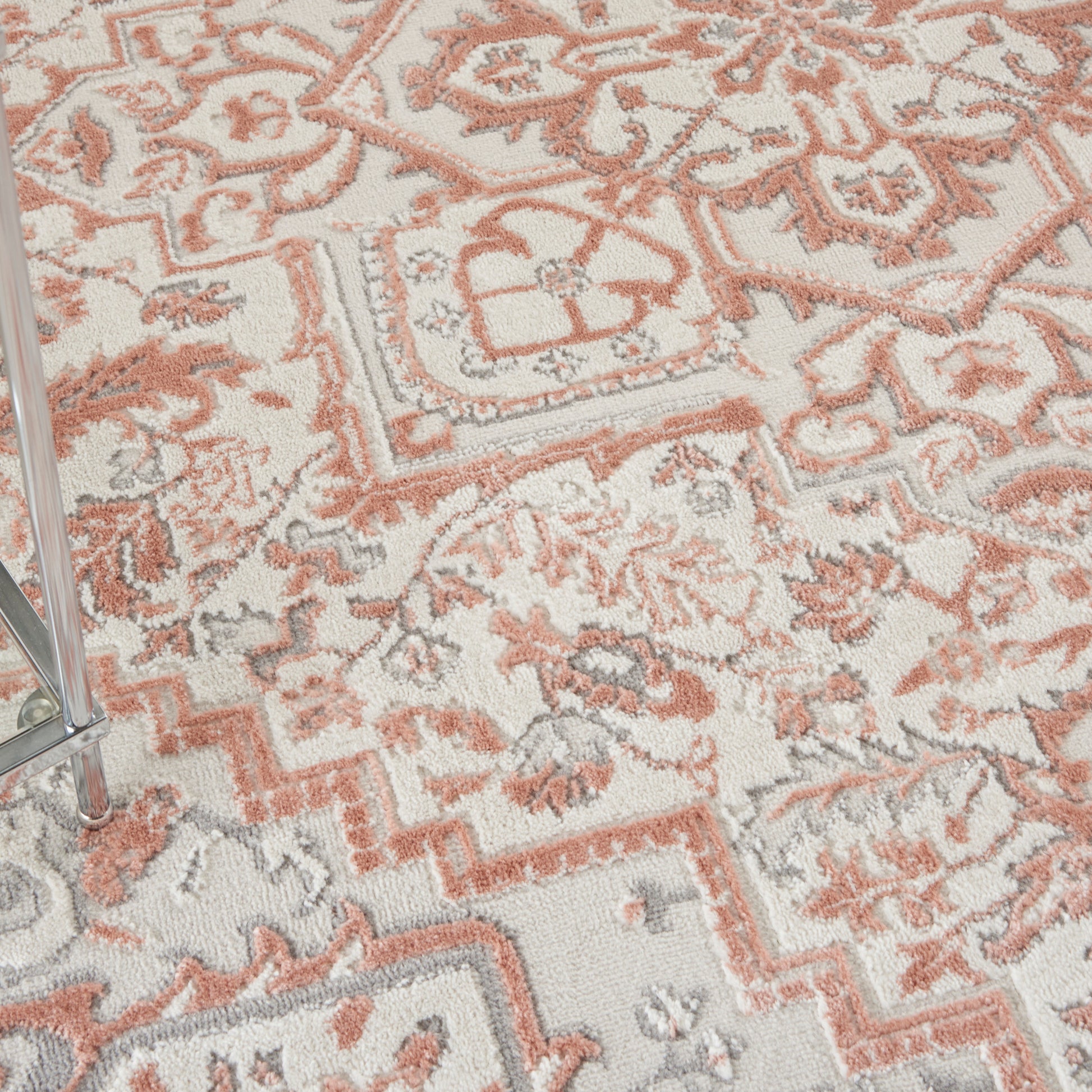 Soft, textured rug with peach and light gray floral and geometric pattern on ivory background; metal chair leg at edge.
