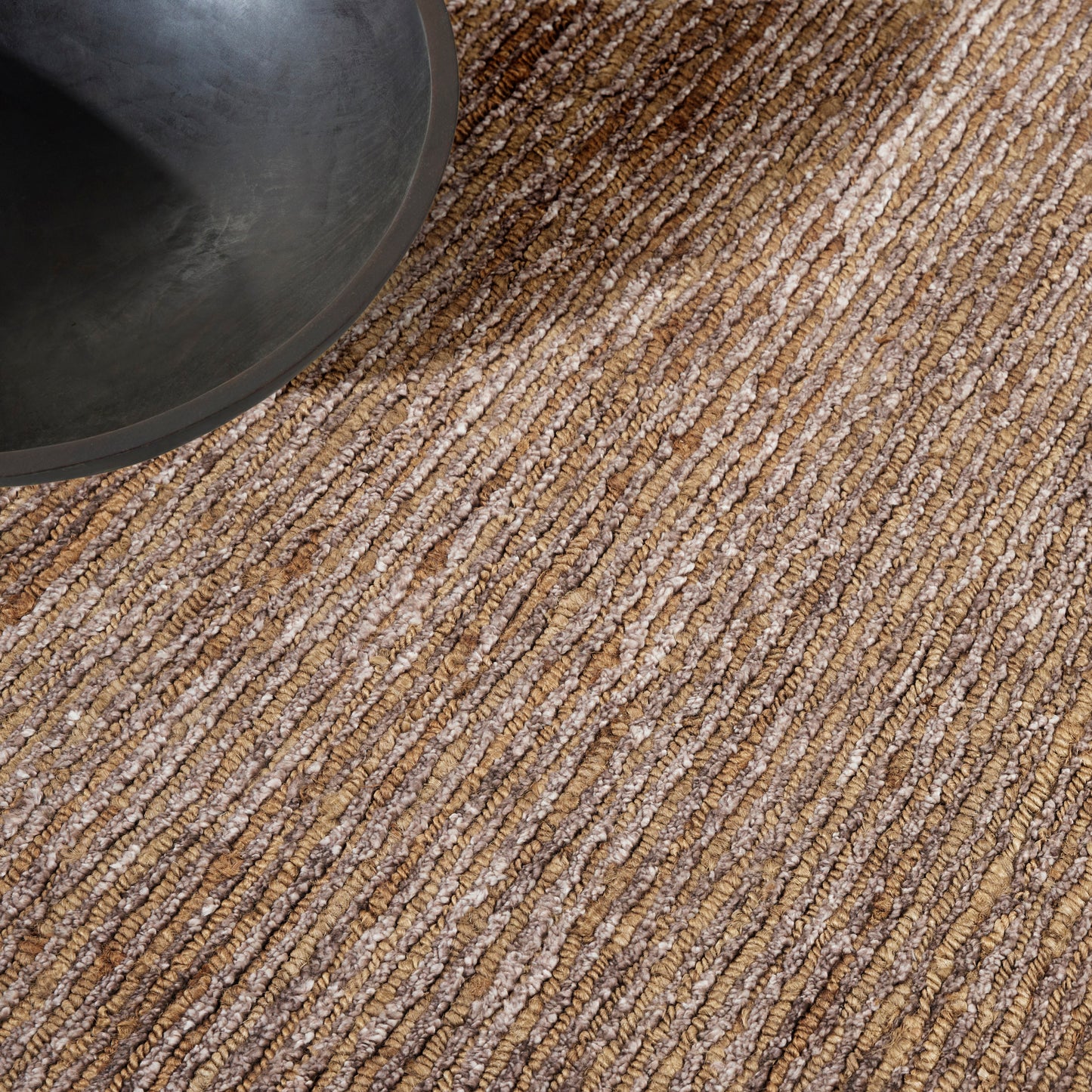 Beige-brown striped looped-jute rug with chunky texture, a dark bowl resting on it.