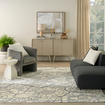 Neutral living room with light wood sideboard, abstract art, ceramic lamp, candles, gray armchairs, dark sofa, beige curtains, patterned rug.