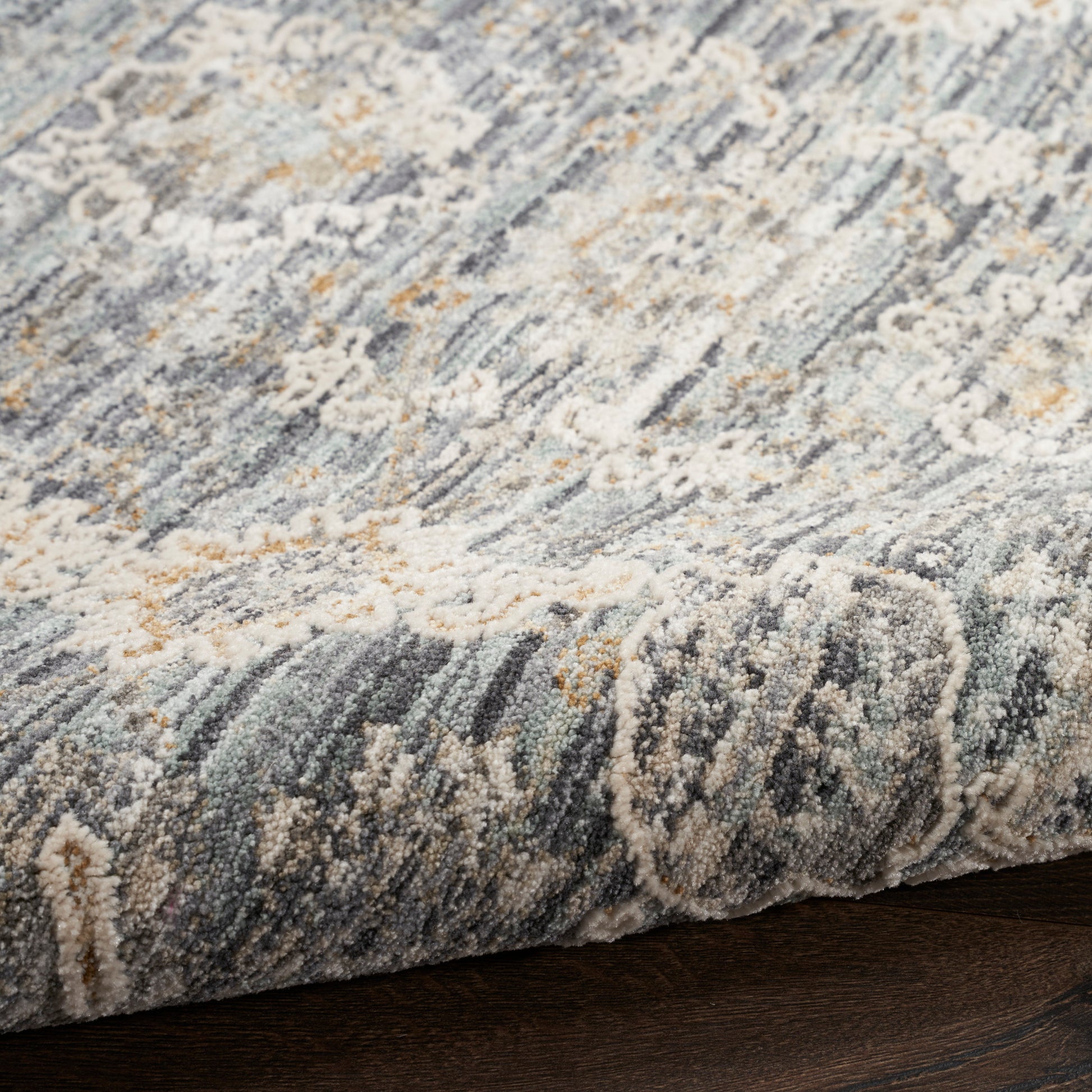 Blue-gray, ivory & tan abstract rug with soft tufted pile, edge curling over dark wood floor.