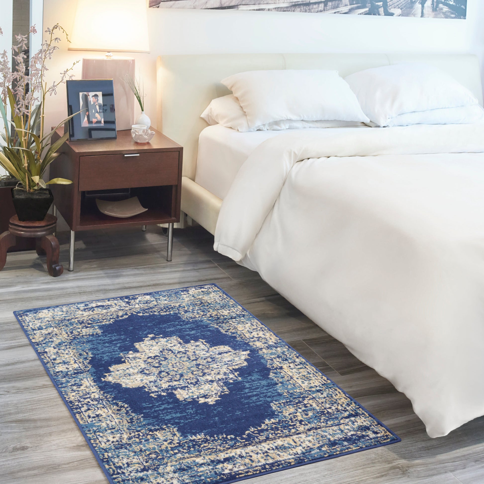 Rectangular blue distressed rug with cream medallion pattern on gray wood floor beside white bed and dark wood nightstand.
