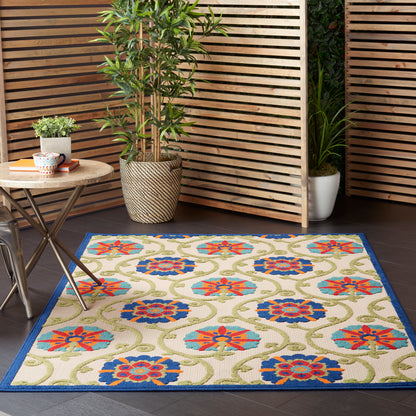 Outdoor patio rug with blue border & colorful floral pattern on cream, beside wooden slat screens & potted plants.