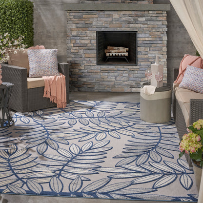Outdoor lounge with stacked stone fireplace, wicker seating, blue leaf rug, pink throws, lanterns and potted plants.