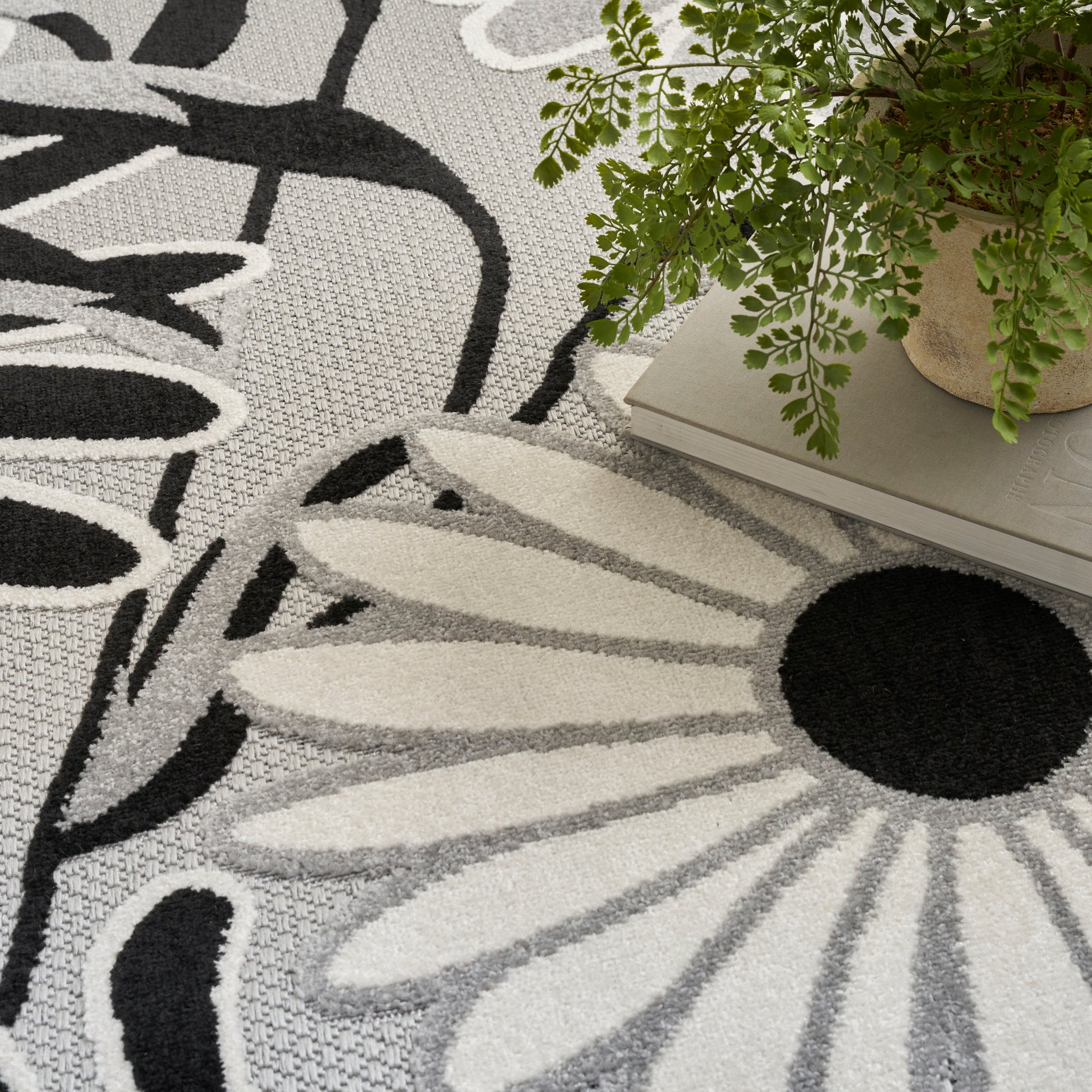 Woven gray daisy-pattern rug with black center and white petals; potted fern sits on books nearby.
