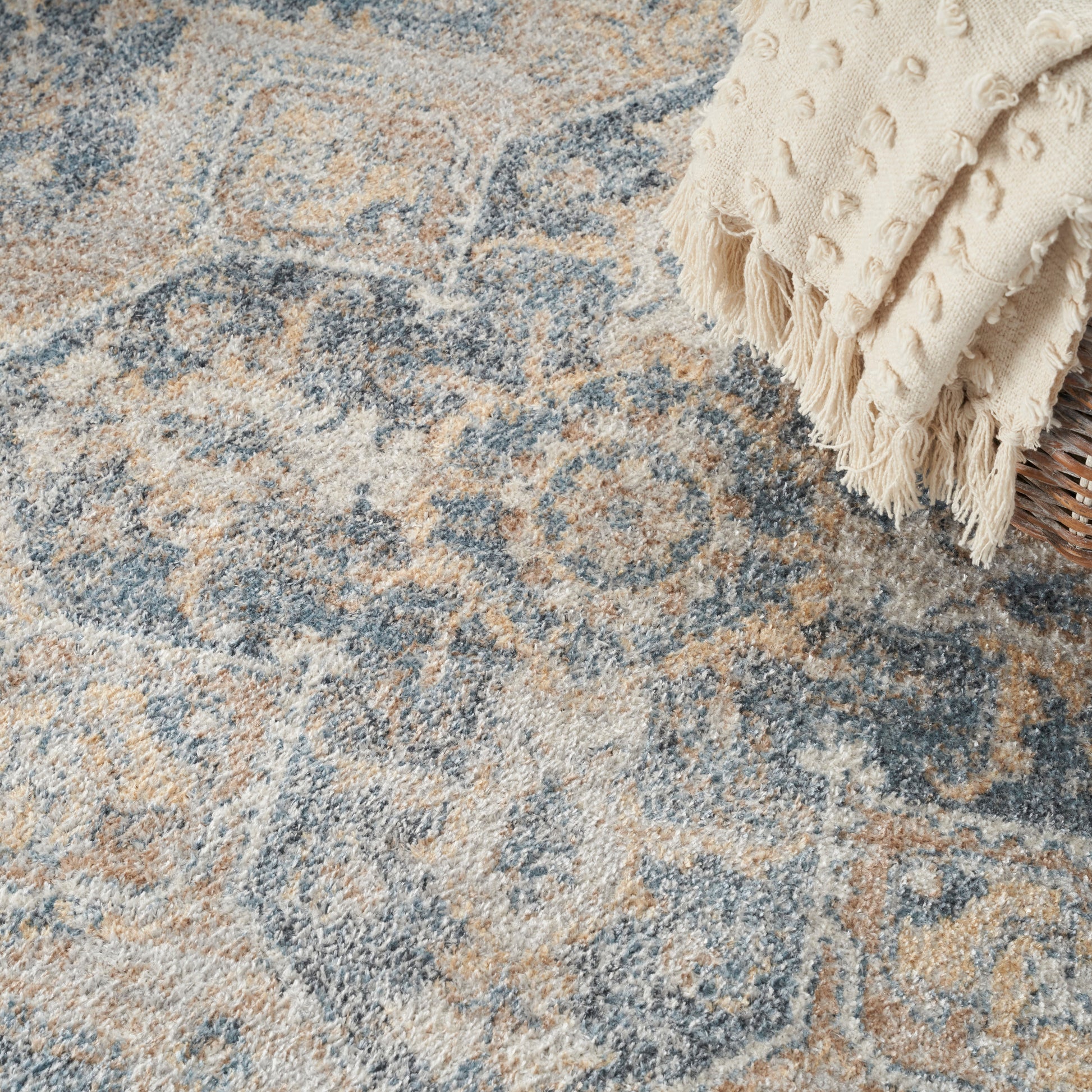 Close-up of a blue, beige and tan patterned oriental rug with a cream fringed throw draped in the corner.