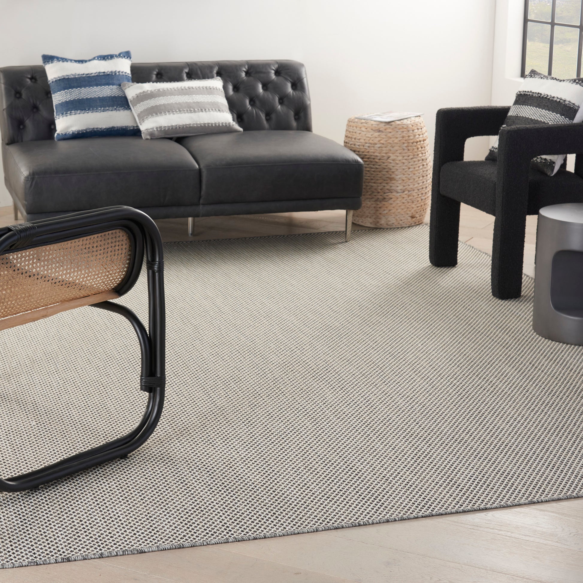 Modern living room with black tufted sofa, striped blue and gray pillows, cane chair, woven pouf, and gray rug.