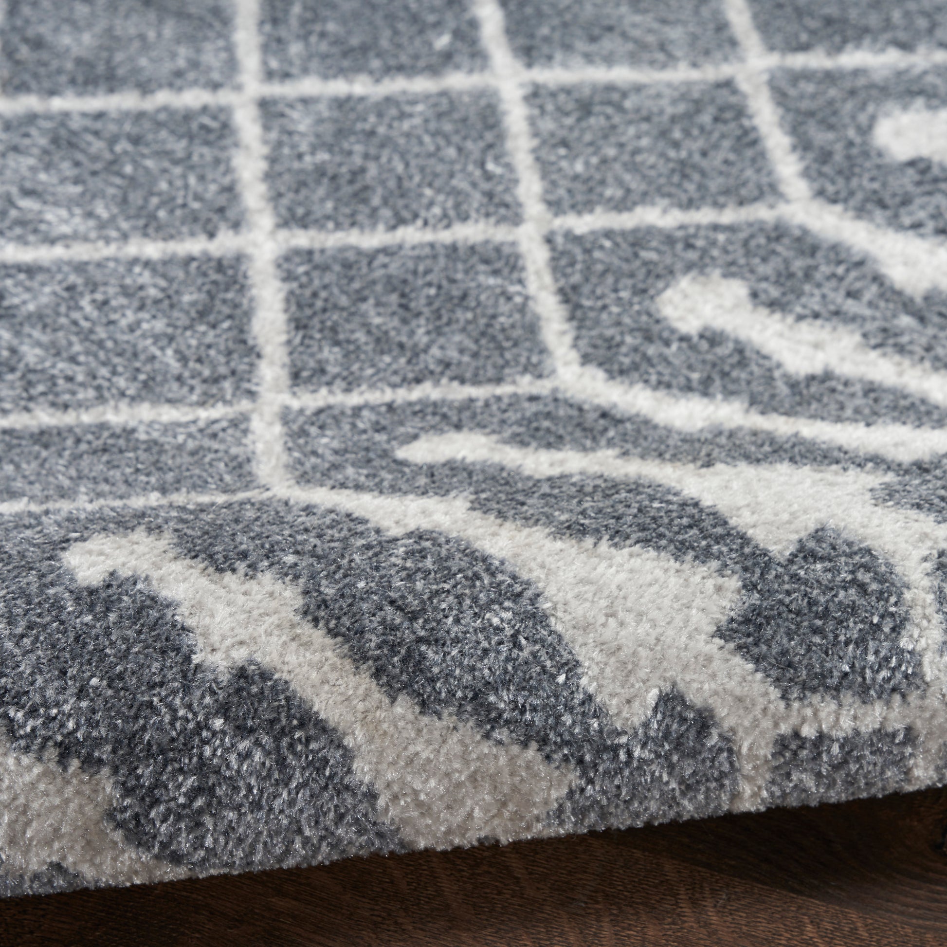 Close-up of a grey and ivory rug with geometric lattice pattern, plush pile, edge visible, dark wood floor.