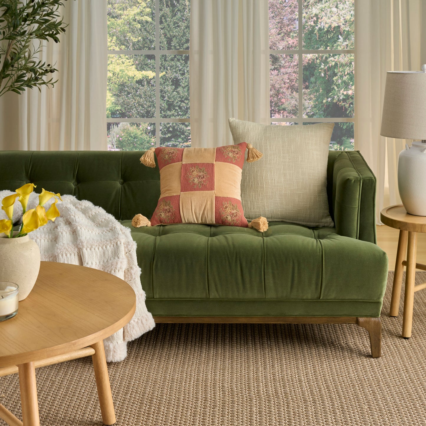Olive green velvet tufted sofa with beige and patchwork cushions, round wooden coffee table, plant, lamp, woven rug.