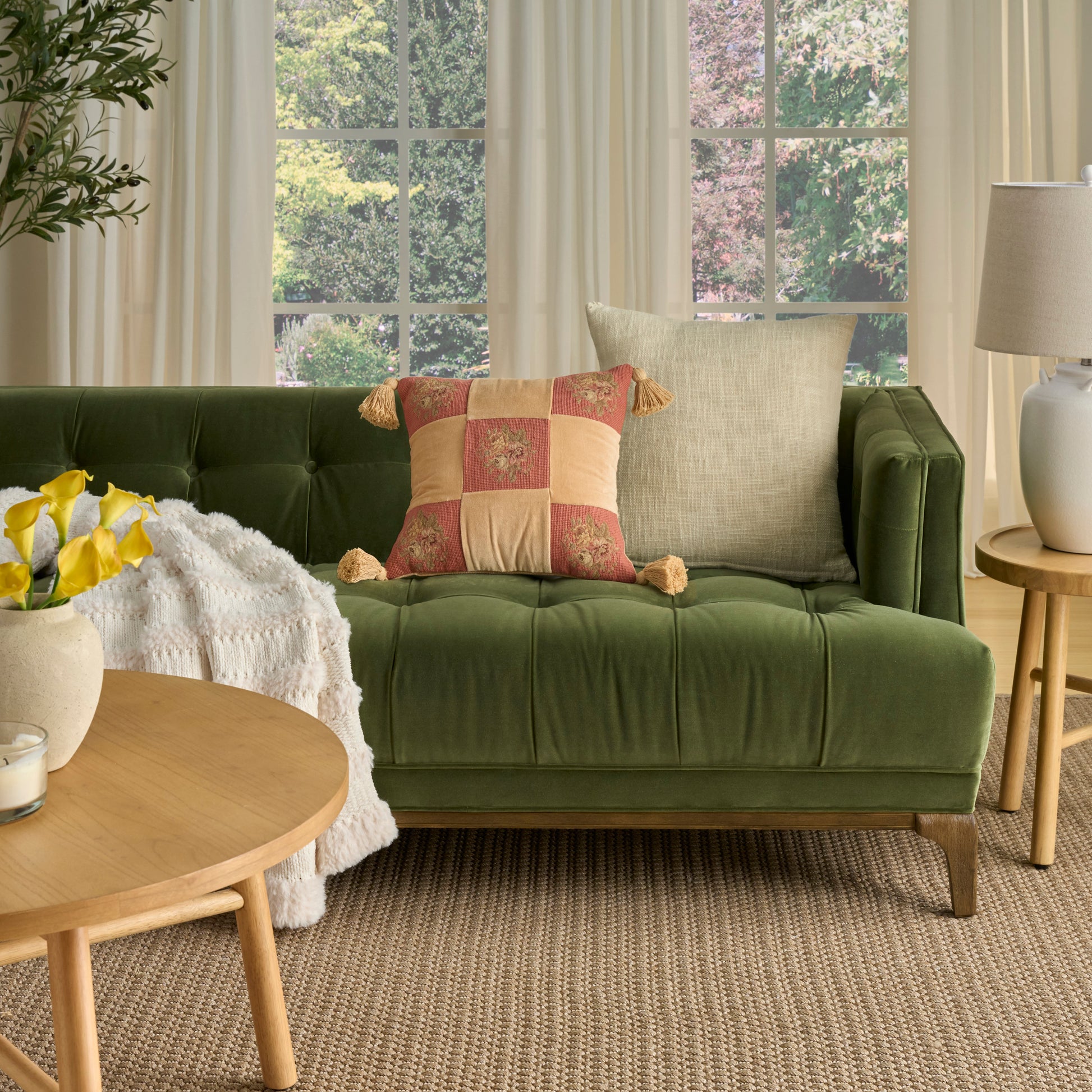 Olive green velvet tufted sofa with beige and patchwork cushions, round wooden coffee table, plant, lamp, woven rug.