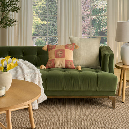 Olive green velvet tufted sofa with beige and patchwork cushions, round wooden coffee table, plant, lamp, woven rug.