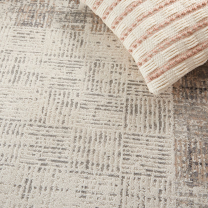 Beige and gray textured area rug with a geometric square-grid pattern; folded cream-pink striped throw at the corner.