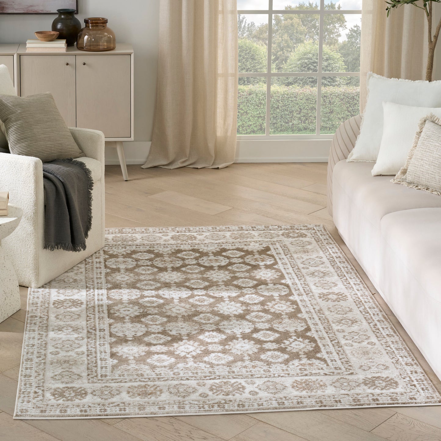 Beige ornate area rug with cream border on light wood floor in a neutral living room with cream sofa and armchair.