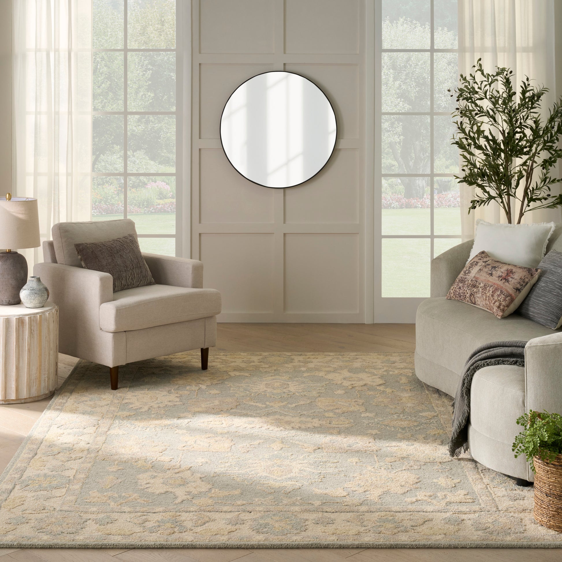 Neutral living room with beige sofa & armchair, round wall mirror, pale patterned rug, and potted plant.