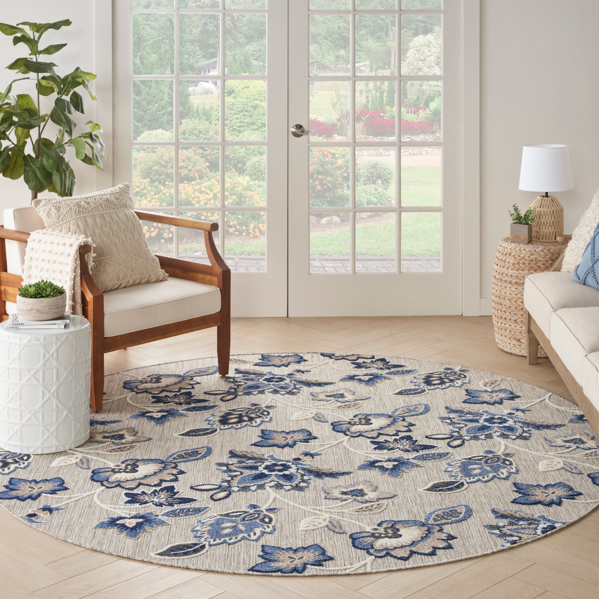 Round beige rug with blue floral pattern in a bright living room; wood armchair, potted plant, wicker lamp, glass doors.