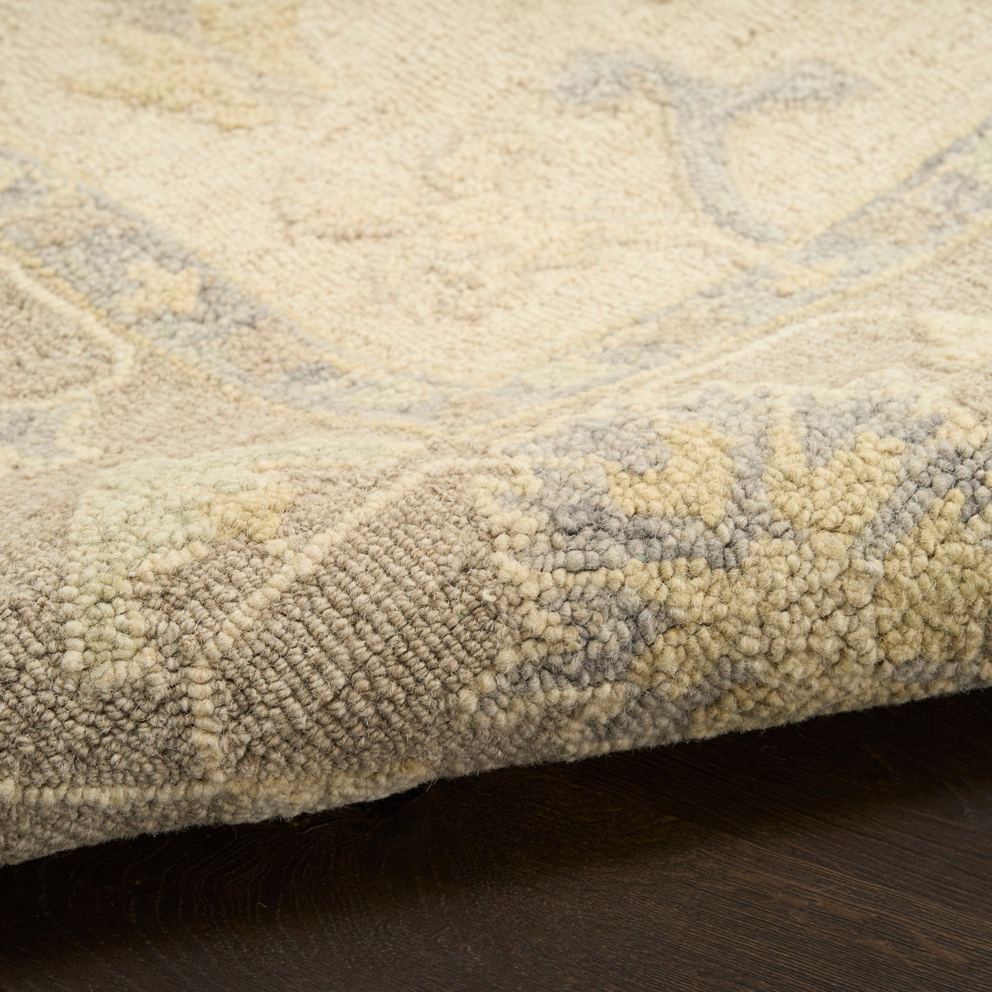 Beige and cream rug with subtle blue-gray pattern; plush high-pile texture and rolled edge on dark wood floor.