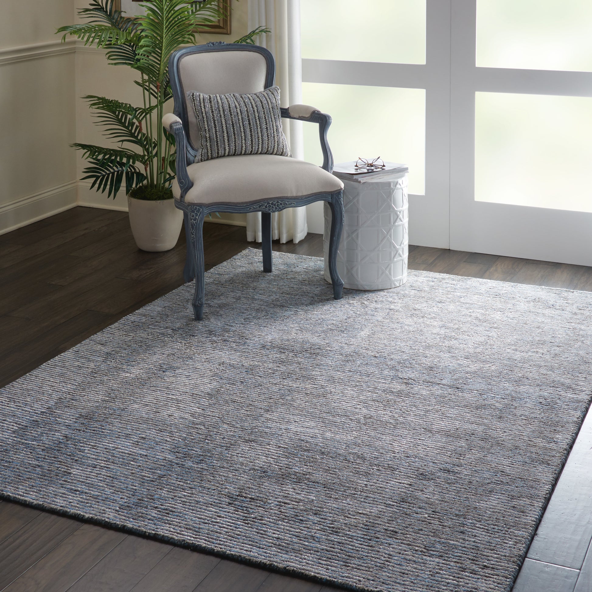 Blue-gray antique French armchair with beige upholstery and striped lumbar cushion, set on a textured gray rug beside a white lattice side table.