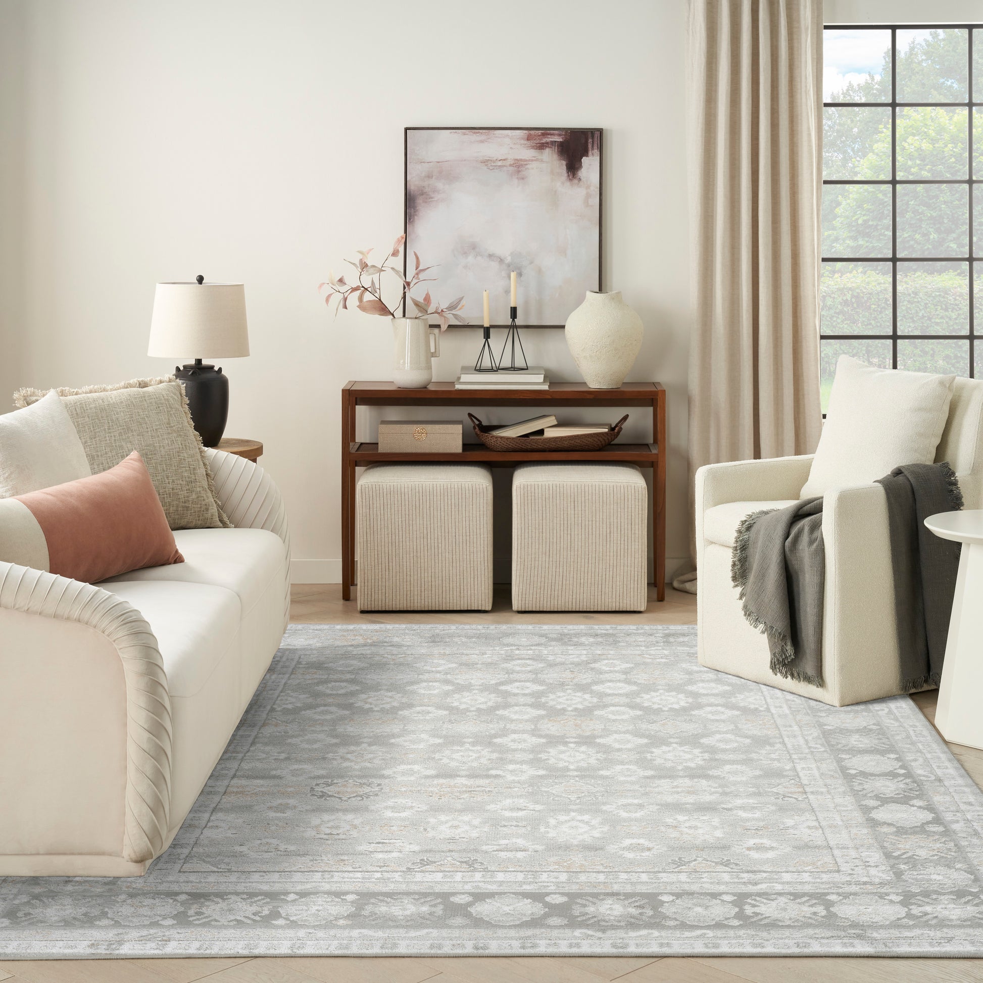 Neutral living room with cream sofa, beige chair, light gray patterned rug, wooden console and two ottomans.