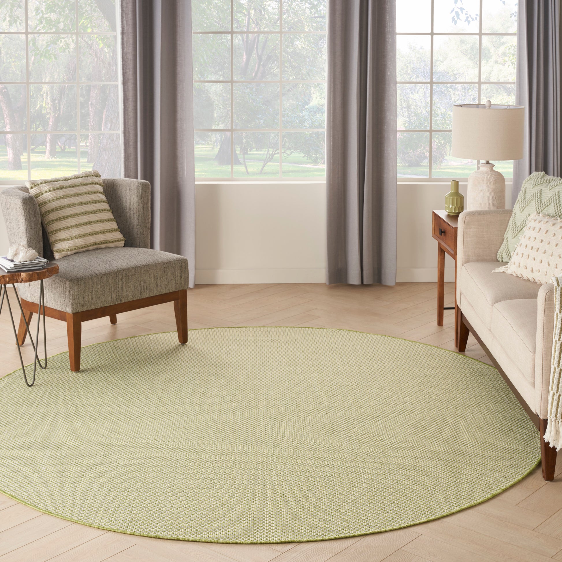 Round olive-green area rug on light wood floor between gray-curtained windows, beige sofas and wooden side tables.