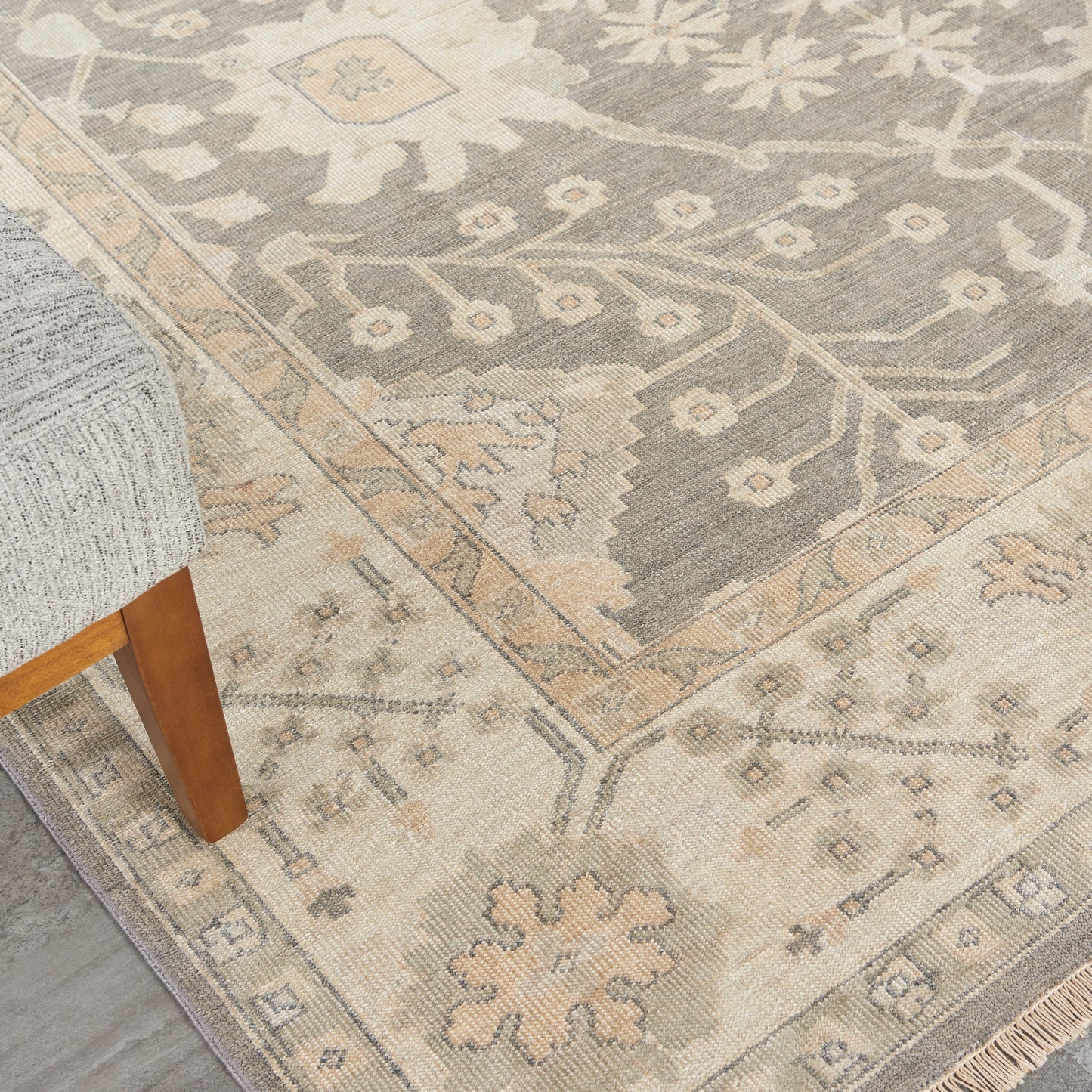 Muted gray-beige Persian-style rug with floral medallion, cream border, fringe edge, wooden sofa leg nearby.