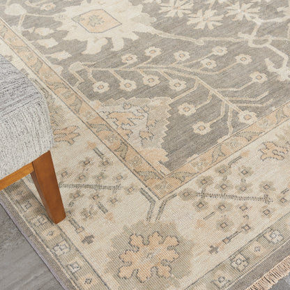 Muted gray-beige Persian-style rug with floral medallion, cream border, fringe edge, wooden sofa leg nearby.