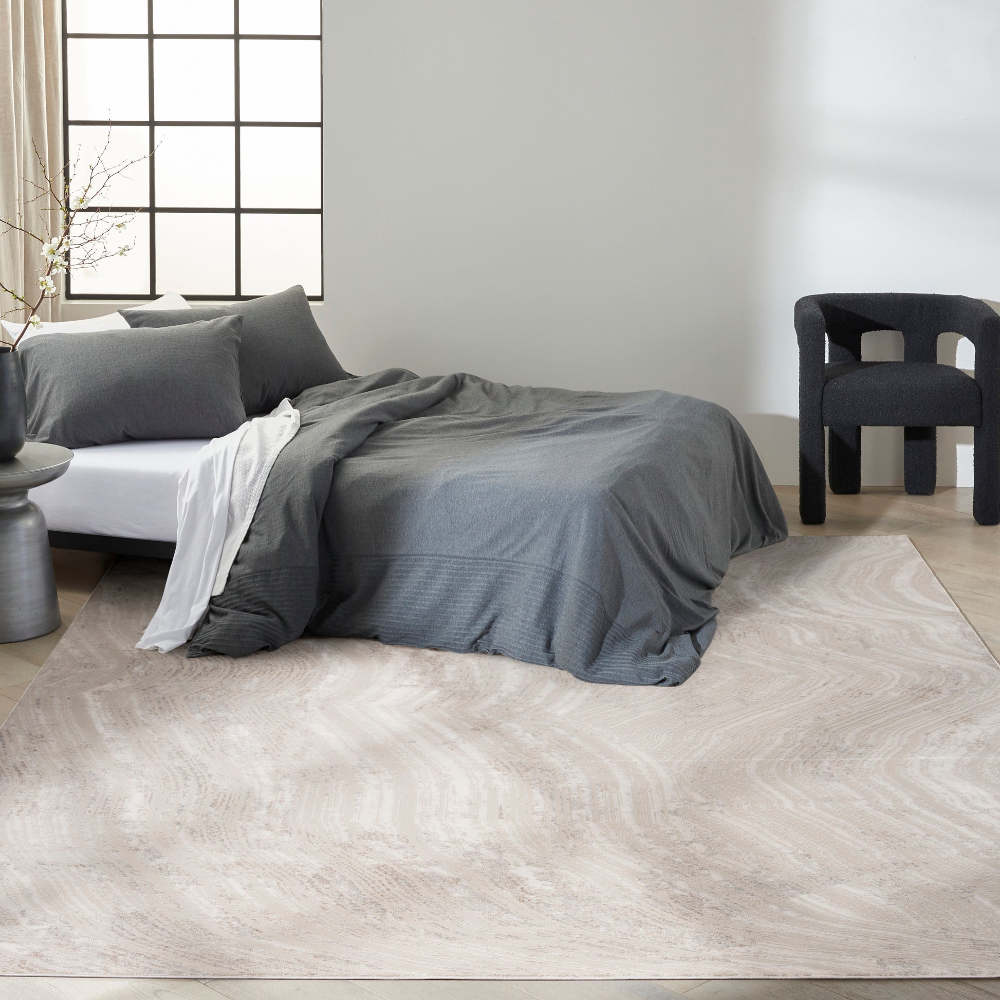 Light beige area rug on a minimalist bedroom floor, with dark grey duvet and pillows on a low bed and a black armchair.