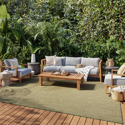 Outdoor lounge set with gray-cushioned wood-framed sofa, chairs & coffee table on a green rug amid lush tropical plants.