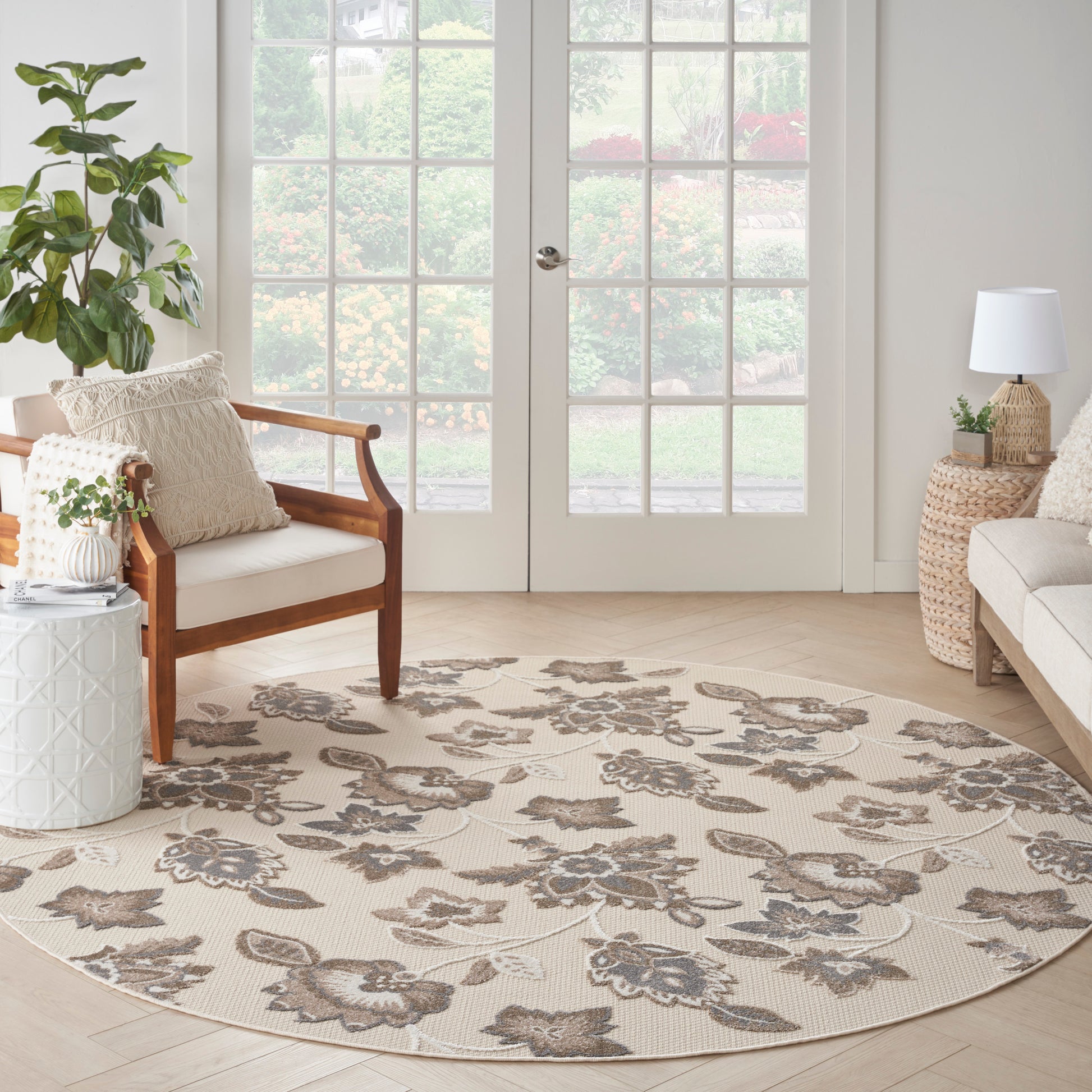 Bright living room with round beige floral area rug, wooden armchair, plants, wicker side tables, and French doors.