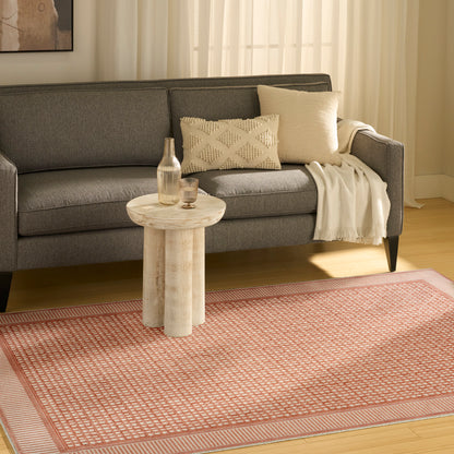 Gray sofa with cream textured pillow and throw, round pale wood side table with bottle and glass, pink patterned rug.