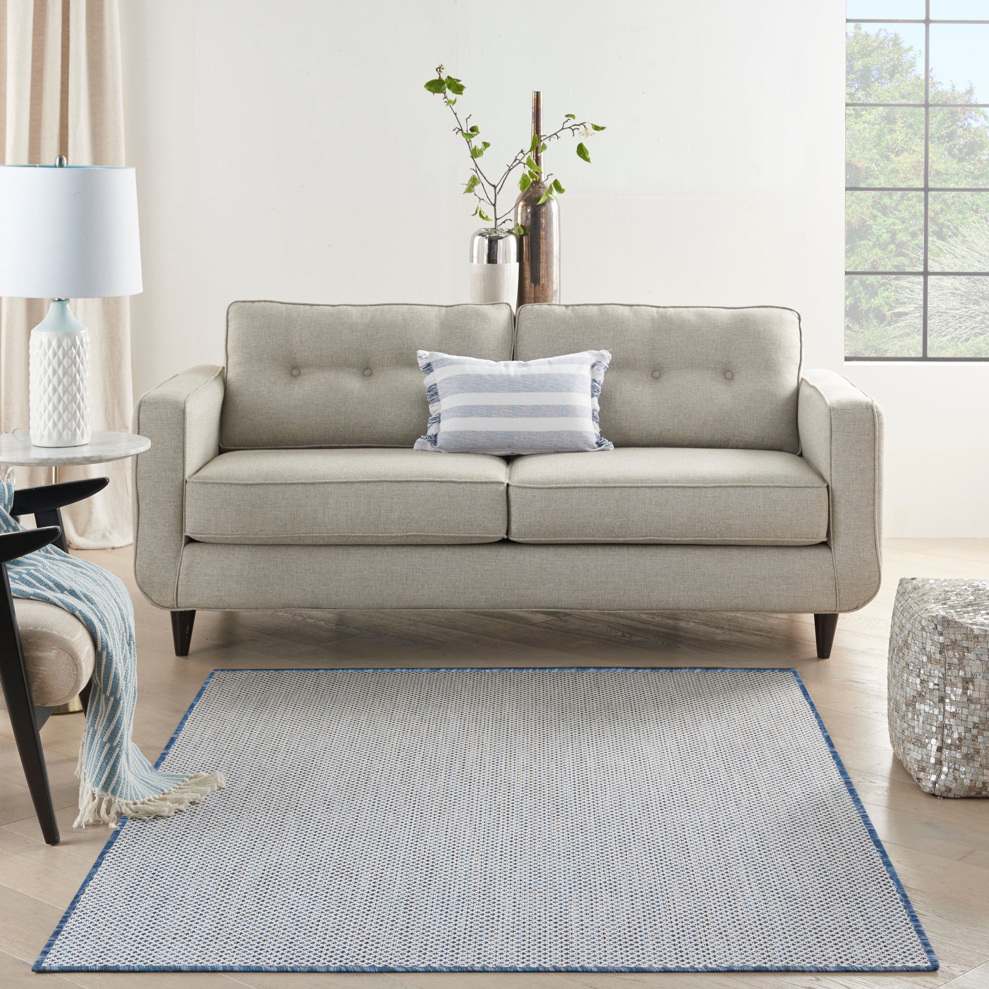 Light gray mid-century sofa with tufted back and dark legs, blue striped pillow, in a room with a blue-edged gray rug.
