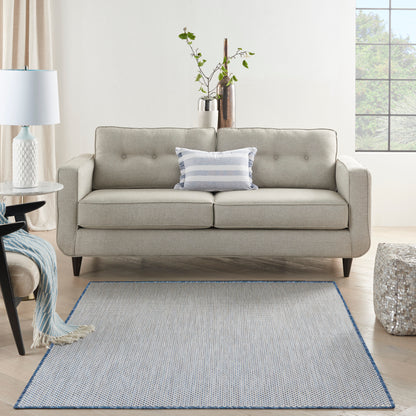 Light gray mid-century sofa with tufted back and dark legs, blue striped pillow, in a room with a blue-edged gray rug.