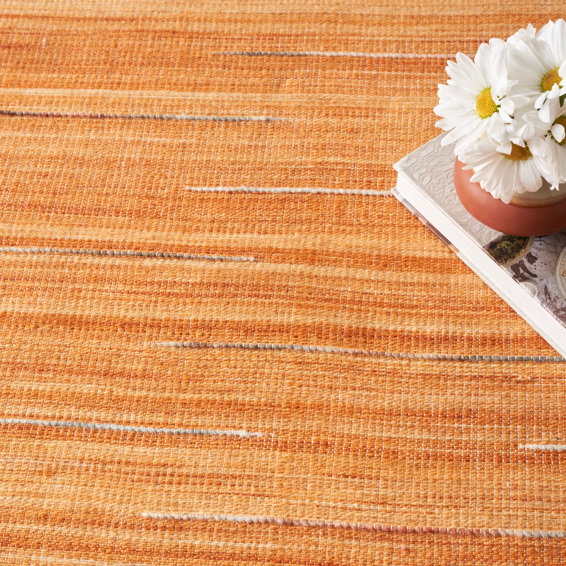 Orange textured rug with subtle stripes; terracotta pot of white daisies rests on a stack of magazines.