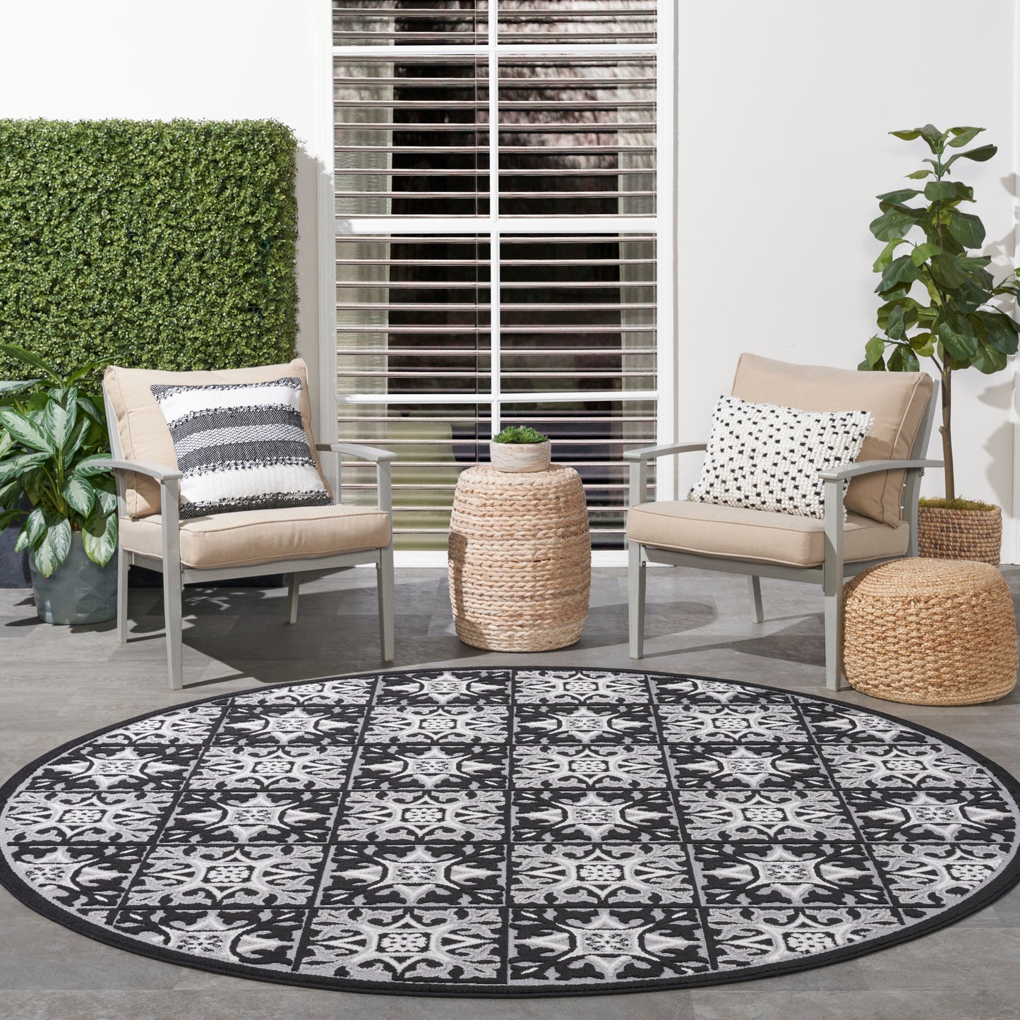 Round black-and-white outdoor rug with intricate pattern on a patio, beige cushioned metal chairs, woven ottomans, and plants.