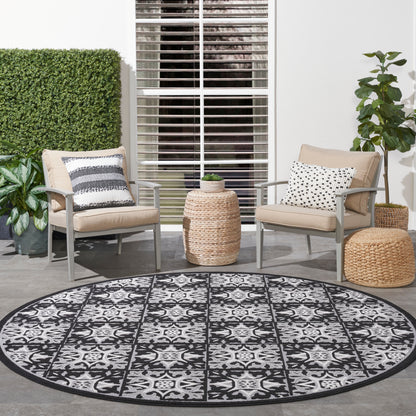 Round black-and-white outdoor rug with intricate pattern on a patio, beige cushioned metal chairs, woven ottomans, and plants.