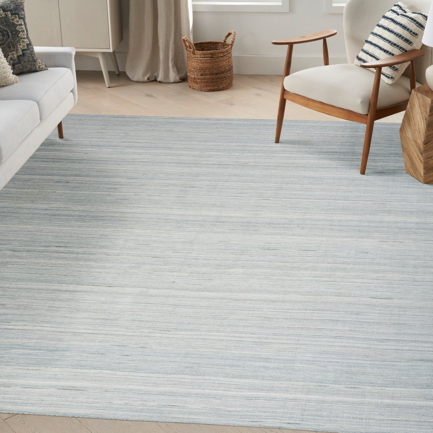 Blue-gray striped rug on pale wood floor with a white sofa, mid-century armchair, and woven basket in a bright living room.