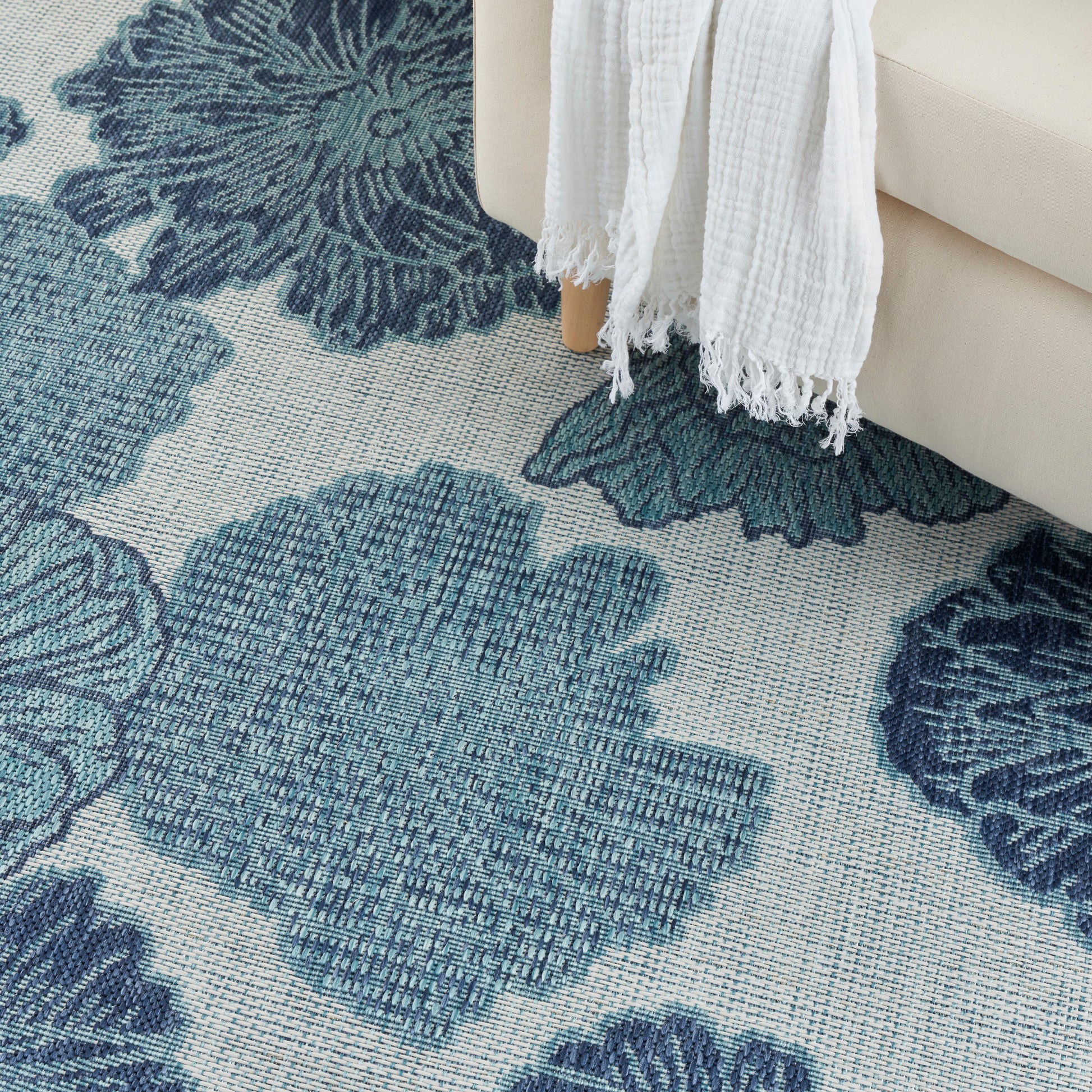 Teal blue floral area rug with large petal motifs, beside a beige sofa with a white fringed throw draped over the edge.