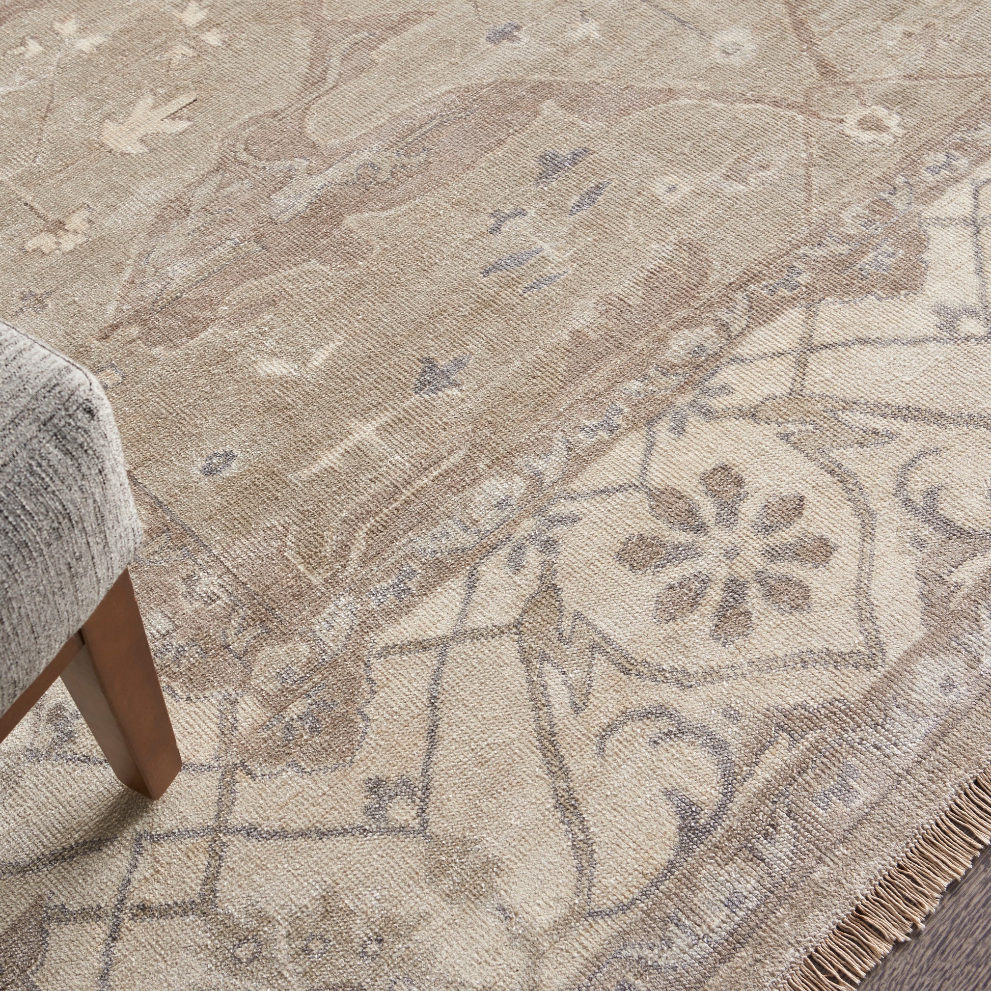 Beige patterned area rug with floral medallion border and fringe, shown with a gray upholstered chair leg.