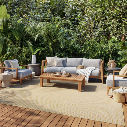 Teak outdoor patio set with light gray cushions: sofa, armchairs, coffee table on a beige rug, tropical garden backdrop.