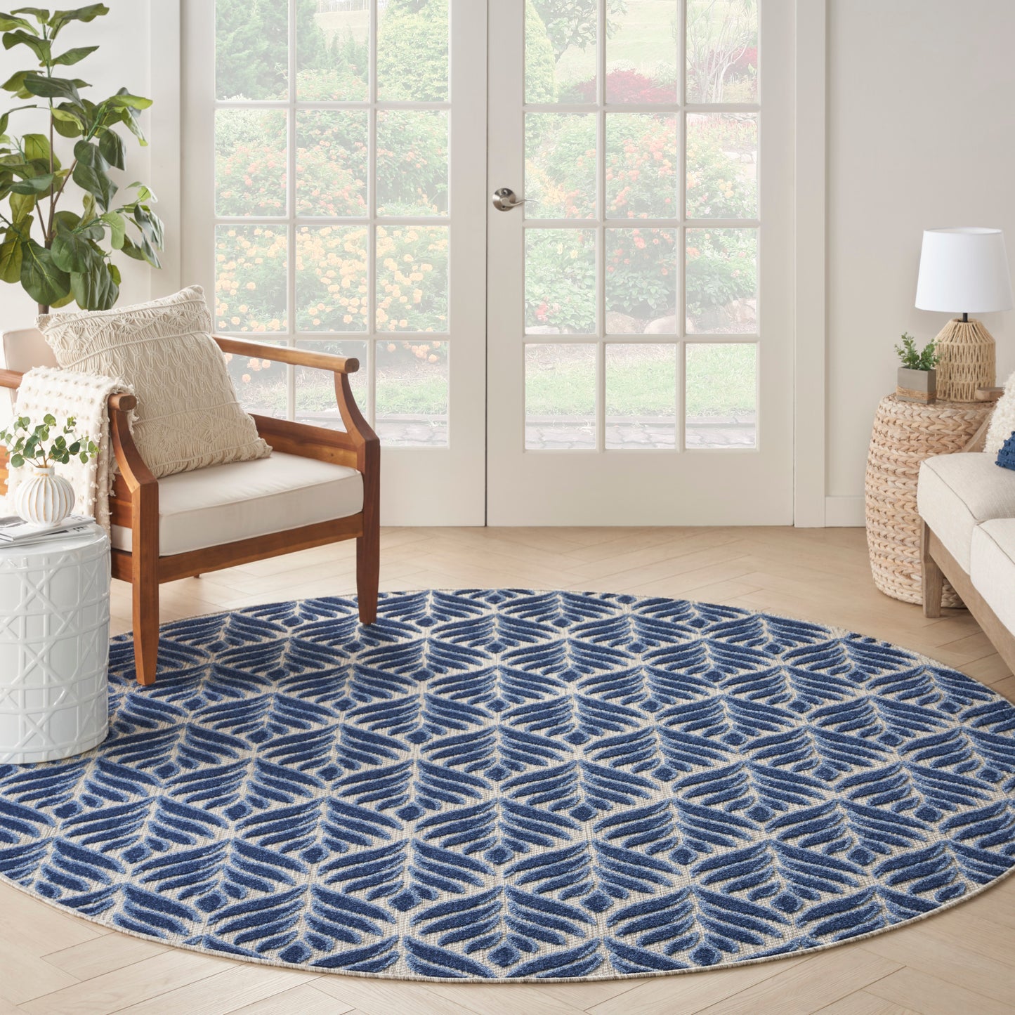 Round blue and white geometric rug on light wood floor in bright living room with wooden armchair, plants, and French doors.